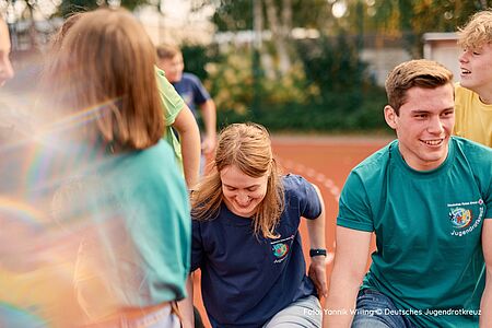 Aktiv im Jugendrotkreuz Betreuer und Kinder des Jugendrotkreuz sitzen im Freien auf einem Sprtplatz